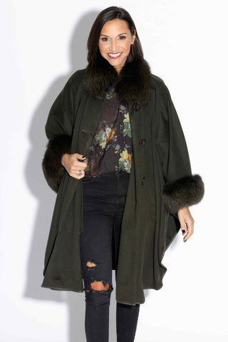 smiling woman wearing an olive cashmere & wool blend cape with fox fur trim against a white backdrop