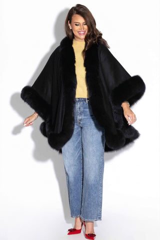 smiling woman wearing a black cashmere blend cape with black fox fur trim against a white backdrop
