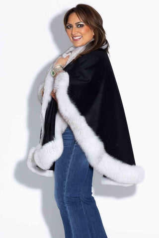 side profile of smiling woman wearing a black cashmere blend cape with white fox fur trim against a white backdrop