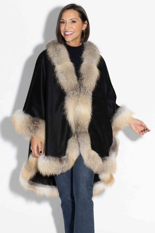 smiling woman wearing a black cashmere blend cape with golden island fox fur trim against a white backdrop