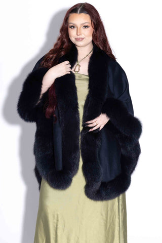 smiling woman wearing a navy cashmere blend cape with fox fur trim against a white backdrop