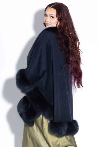 side profile of woman wearing a navy cashmere blend cape with fox fur trim against a white backdrop