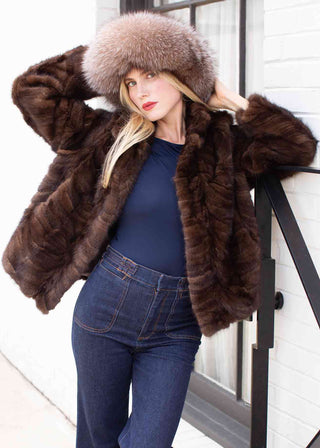 lady wearing a fur hat and brown sectioned mink fur jacket with notch collar, leaning on a gate