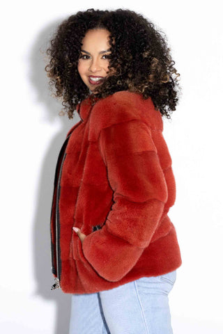 side profile of woman wearing a paprika mink fur jacket with horizontal pattern and zip front, with her hands in the pockets, against a white backdrop