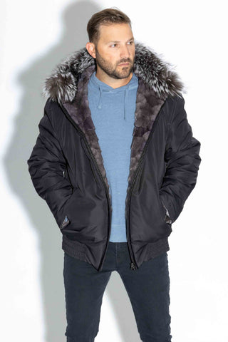 man wearing a reversible blue iris sectioned mink fur bomber jacket with silver fox fur trimmed hood, with the black cloth side facing out, against a white backdrop