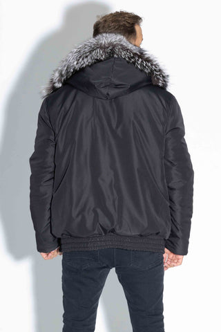 back of a man wearing a reversible blue iris sectioned mink fur bomber jacket with silver fox fur trimmed hood, with the black cloth side facing out, against a white backdrop