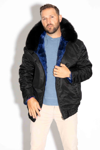 man wearing a reversible blue cross sectioned mink fur bomber jacket with black fox fur trimmed hood, with the black cloth side facing out, against a white backdrop
