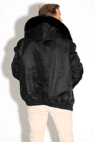 back of a man wearing a reversible blue cross sectioned mink fur bomber jacket with black fox fur trimmed hood, with the black cloth side facing out, standing against a white backdrop