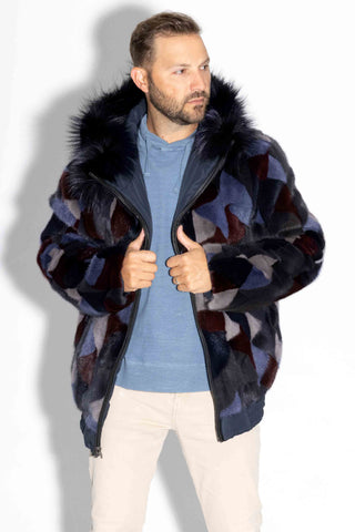 man wearing a reversible blue multicolor sectioned mink fur bomber jacket with navy fox fur trimmed hood against a white backdrop