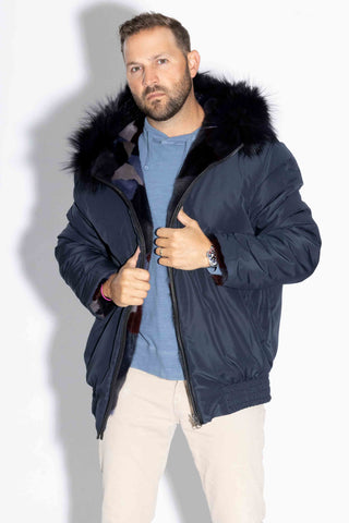 man wearing a reversible blue multicolor sectioned mink fur bomber jacket with navy fox fur trimmed hood, with the navy cloth side facing out, against a white backdrop