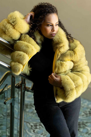 woman wearing a yellow fox fur jacket with horizontal pattern and zip front, standing on a staircase