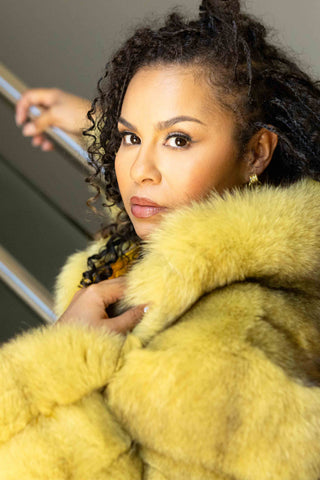 woman wearing a yellow fox fur bolero jacket on a stairwell, against a gray background